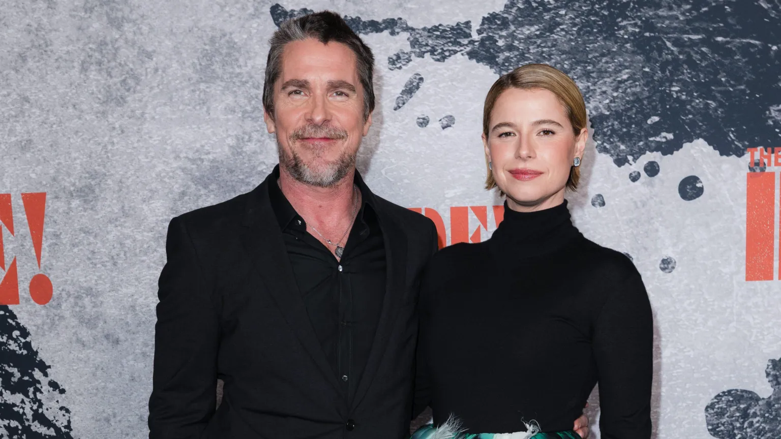  Bale on the left, Buckley on the right. Bale wears a silver chain, and black shirt and jacket. Jessie has jaw-length blonde hair slicked back. She wears a black high neck jumper, with the top of a blue and white skirt visible.