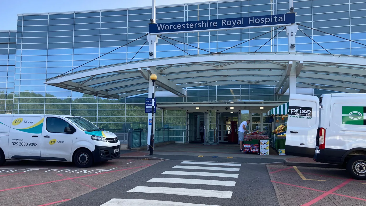 The outside of Worcestershire Royal Hospital. There is a zebra crossing leading to the entrance with an arched metal roof above. A blue sign with white writing shows the hospital's name.