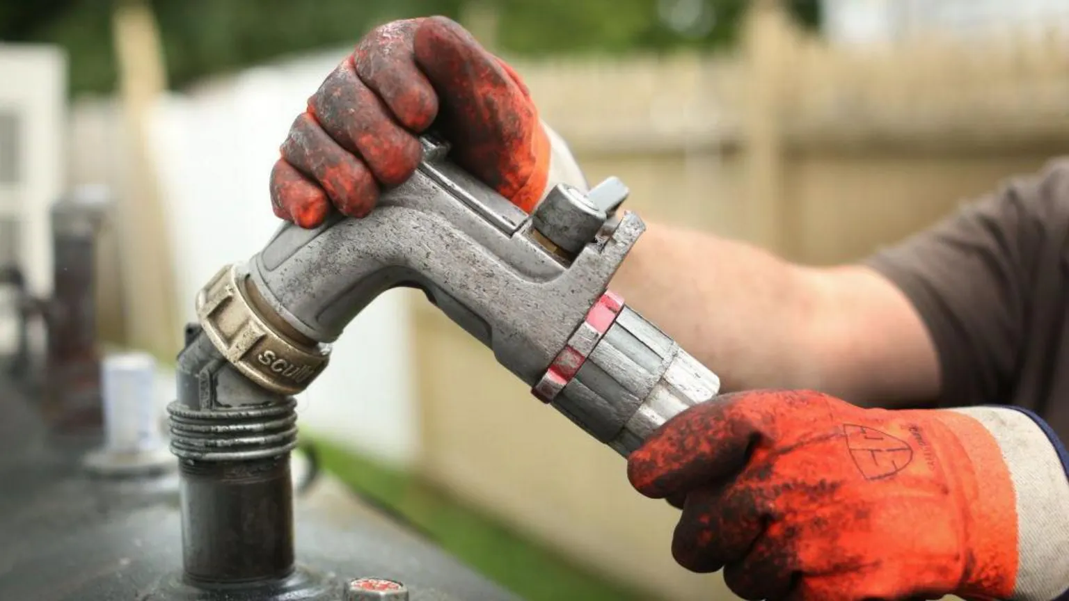 A person with dirty orange gloves holds onto to a metal hose connected to a heating oil tank.