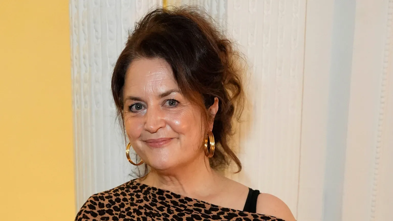  Close-up of Ruth Jones. She wears her brown hair up. She has gold hoops, and wears an off-shoulder leopard print dress.