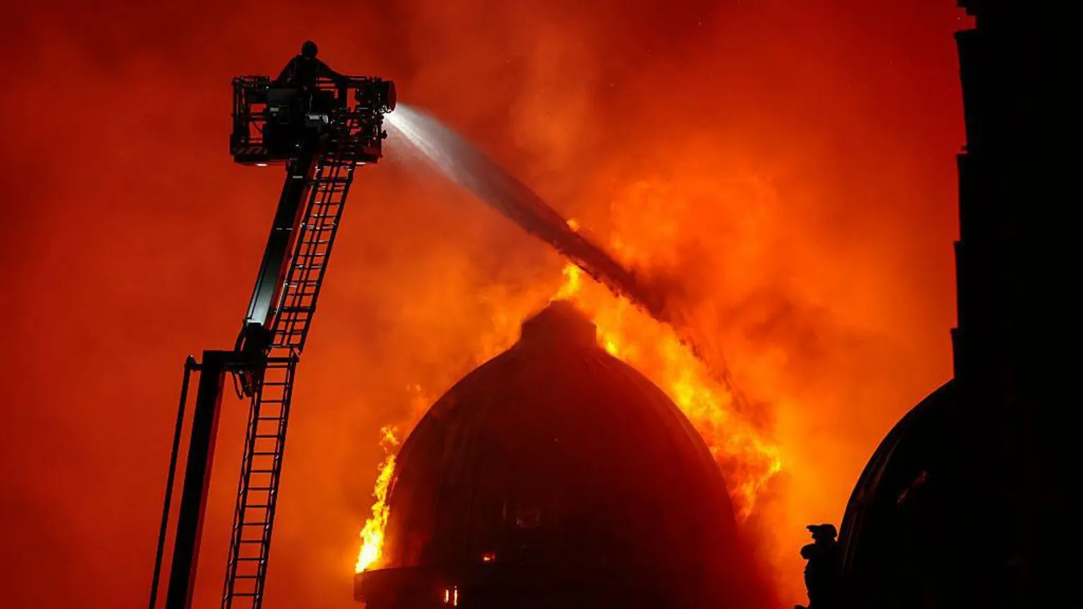 A large domed building on fire. A firefighter on an extendable ladder is spraying water over the flames