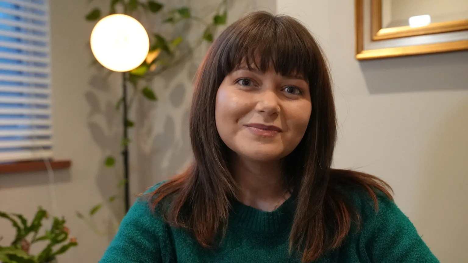 Ann Gannon/BBC A woman wearing a dark green knit sweater is seated indoors. She has straight, medium‑length brown hair with lighter ends. In the background, a warm round lamp glows beside a leafy plant. A window with horizontal blinds is visible on the left, and a framed mirror hangs on the wall to the right.
