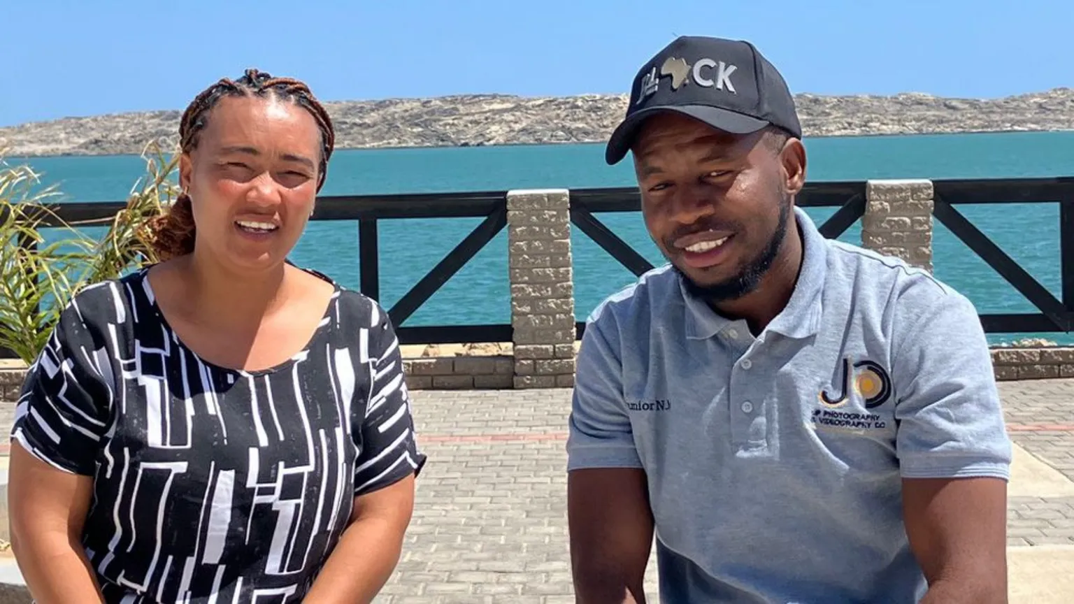 Johannes Dell/BBC Luciel Adams (L) and Junior Mutaleni (R) - two youth activists smile for the camera as they sit by the sea in Lüderitz 