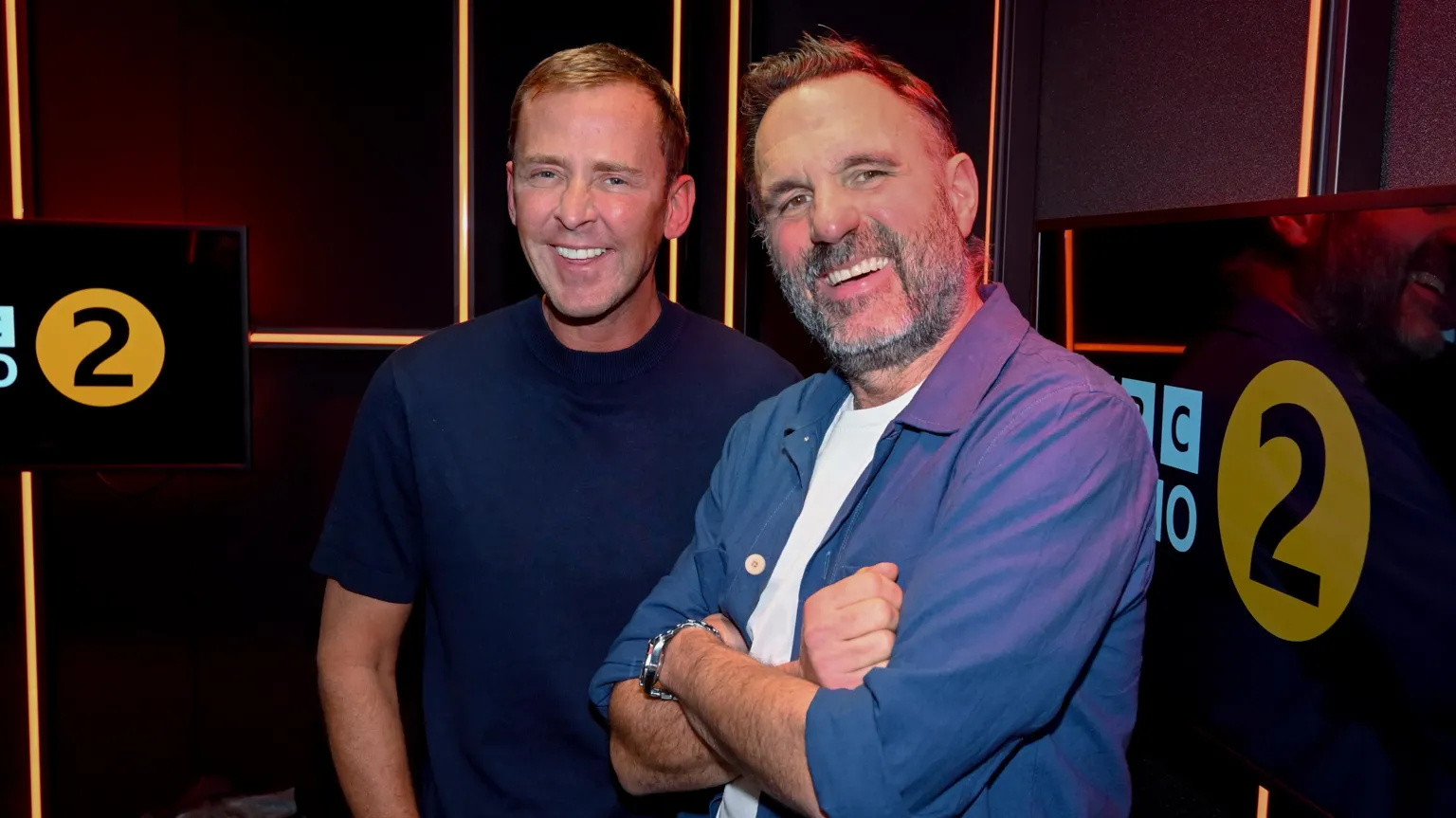 Shaun Keaveny, wearing an open blue collared shirt and white tee, smiles next to Scott Mills, wearing a navy tee.