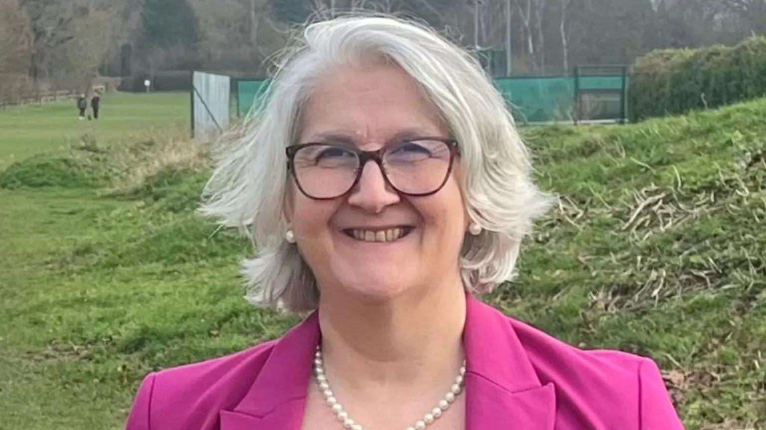 Edna Murphy Edna Murphy is standing in front of a field. She has shoulder length grey hair and is wearing brown framed glasses, pearl earrings and a pink coat. 