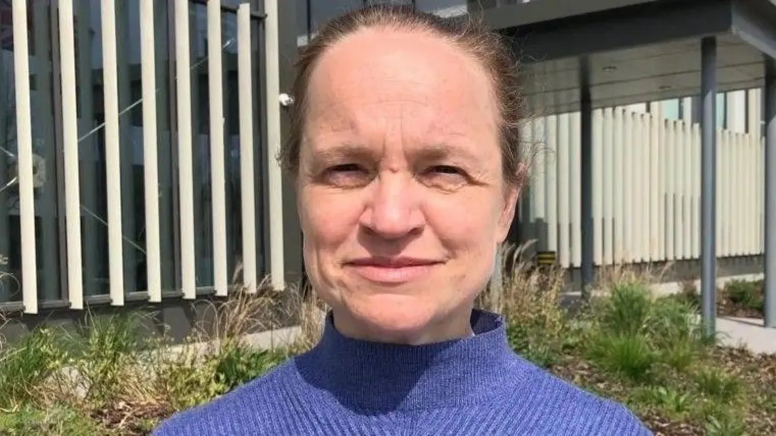 Bryony Goodliffe is standing in front of a modern building which has an entrance on the right. She has brown hair in a ponytail and is wearing a purple jumper. 
