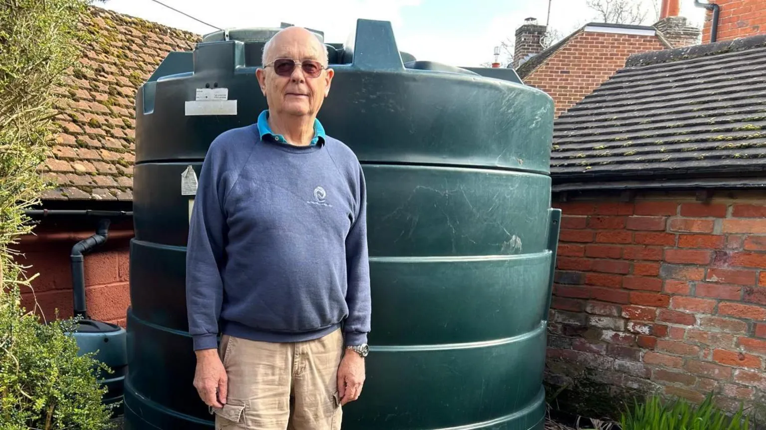 Chris Wheeler Chris Wheeler stands next to his dark green heating oil tank in his garden. He is wearing glasses, a blue sweatshirt and beige cargo trousers. The cylinder-shaped tank is in the corner of the garden in front of an old brick wall.
