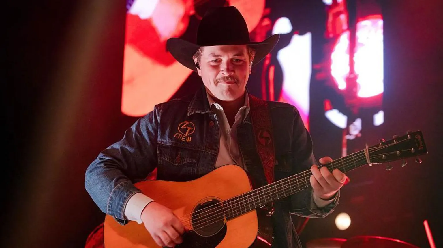  Zach Top wearing a black stetson hat and blue denim jacket plays an acoustic guitar. He is standing on a stage with a red background.