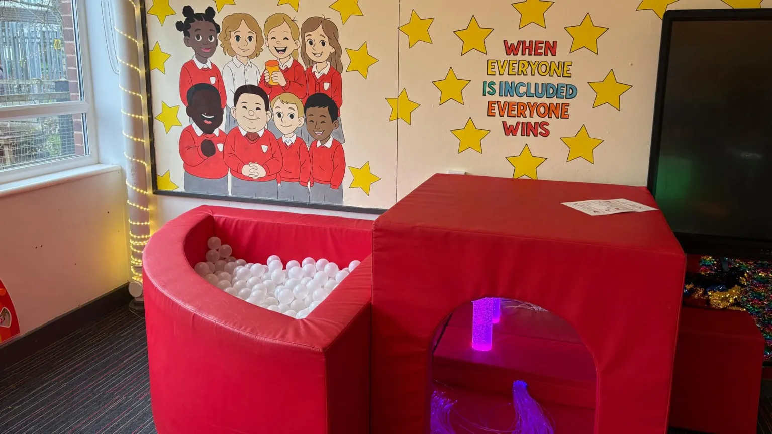 A classroom corner featuring a red soft‑play ball pit filled with white plastic balls next to a red sensory tunnel with purple lighting inside. Behind it is a wall display showing cartoon children in red uniforms surrounded by yellow stars, with the text “WHEN EVERYONE IS INCLUDED EVERYONE WINS.” A large window is on the left with string lights hanging beside it.