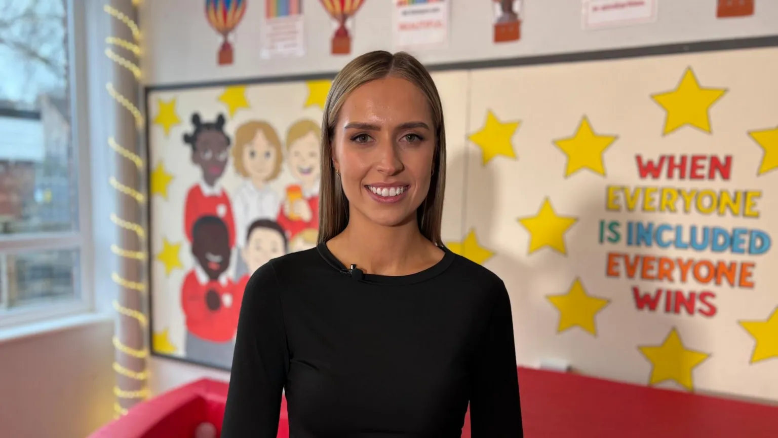 Rachel Donnelly wearing a black top stands in a brightly decorated classroom area. Behind them is a wall display with yellow stars and the slogan “WHEN EVERYONE IS INCLUDED EVERYONE WINS.” To the left are colourful drawings of children in school uniforms, and to the far left is a large window with string lights wrapped around the frame. A red soft‑play structure is partially visible at the bottom of the image.