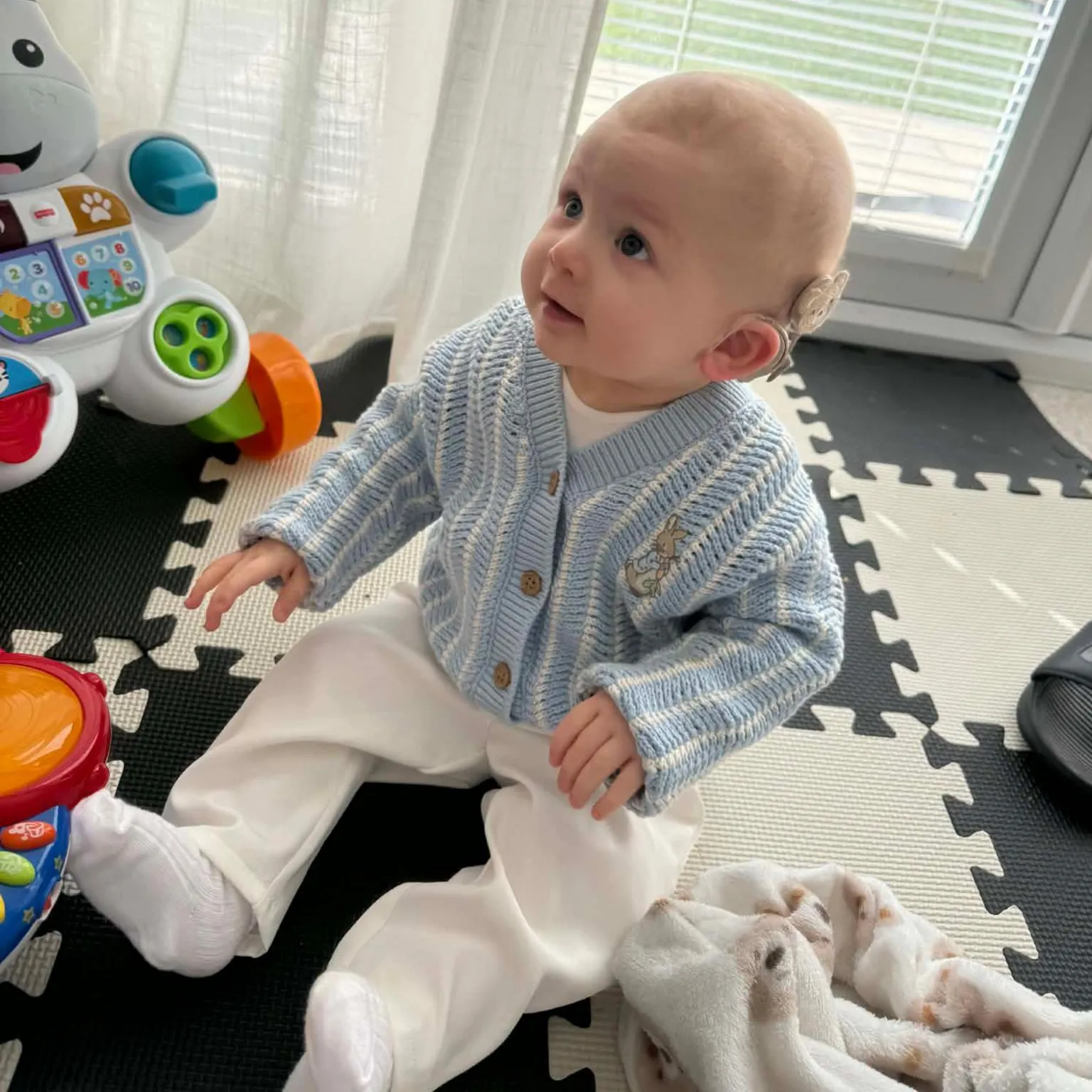 Morgan Deakin A smiling baby wearing a blue cardigan sitting on a black and white play mat