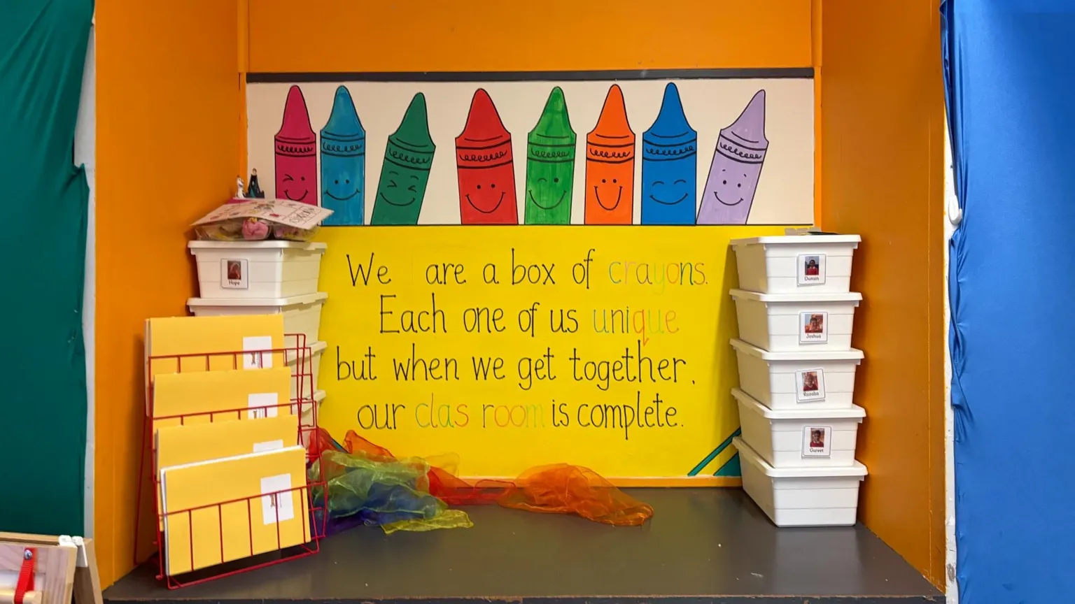 A brightly coloured classroom display shows large cartoon crayons with smiling faces above a yellow sign that reads, “We are a box of crayons. Each one of us unique, but when we get together, our classroom is complete.” On the left are stacked trays and folders in a red rack, and on the right is a tower of white storage boxes. Colourful sheer fabrics are spread out on the floor in front of the display.