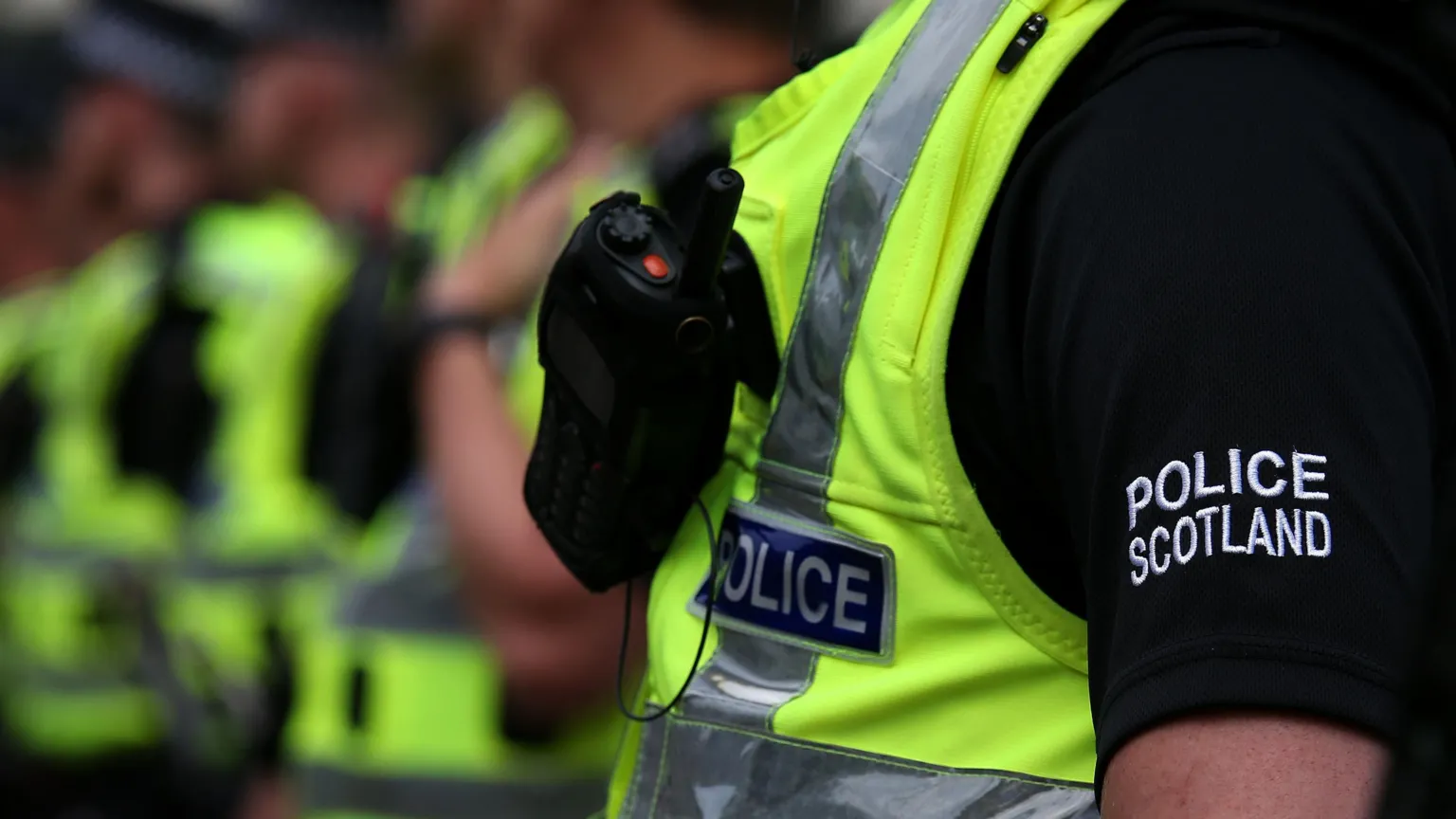  Row of Police Scotland officers, all unidentifiable. It is focused on the Police Scotland embroidery on the officer's black t-shirt. The officers are wearing hi-viz police vests and have radios. 