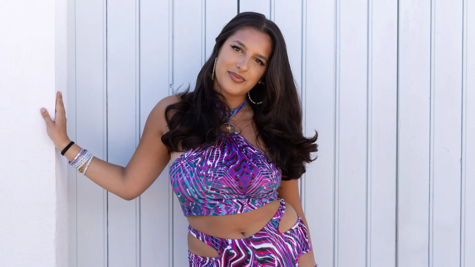 BBC/Twofour Priya has long black hair and wears a bright purple co-ordinated outfit with a cut-out design. She wears bracelts and gold hoops and stands in front of a cream background.