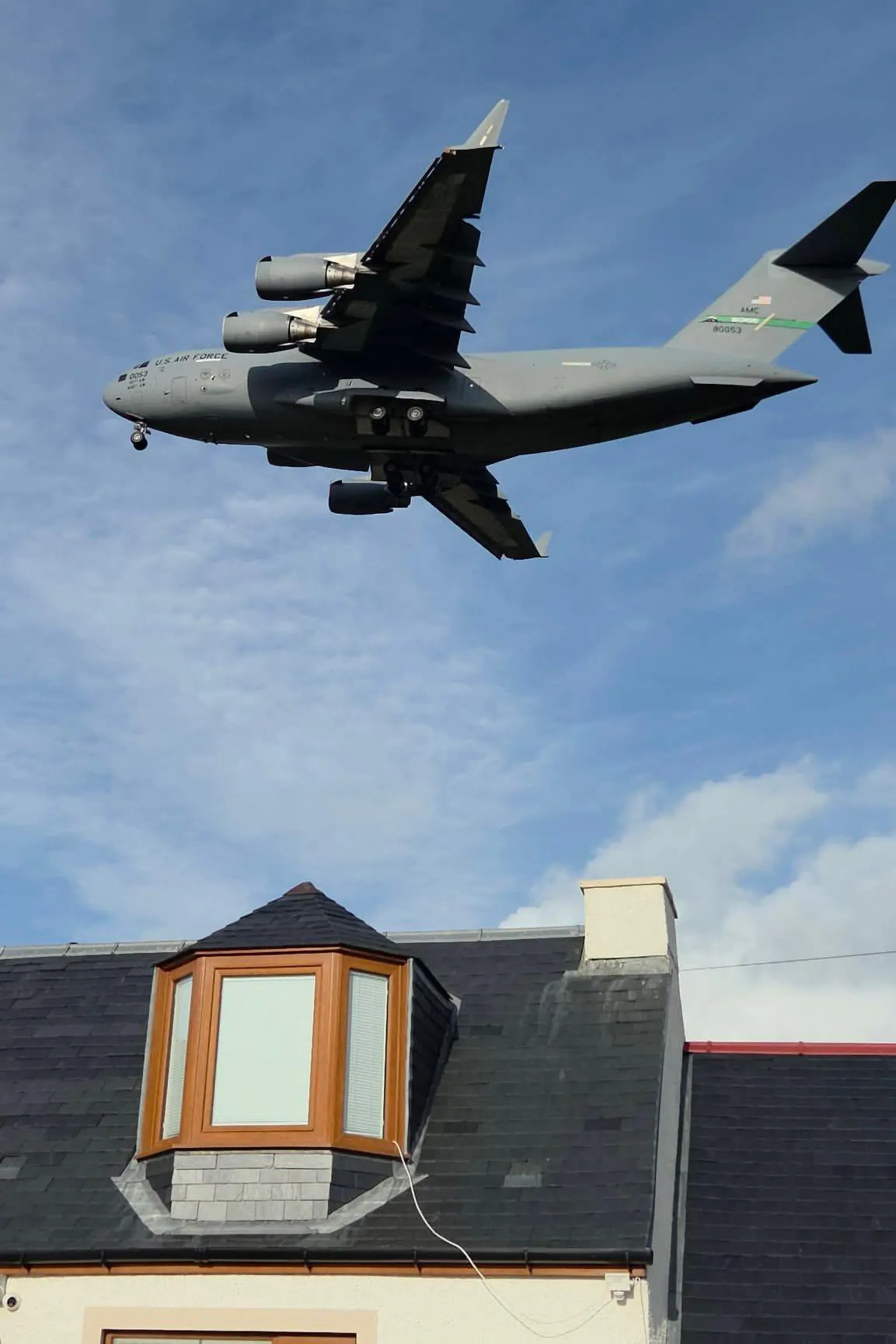Stu Smith A large grey US Air Force military jet aircraft against a blue sky above a house with bay dormer window in the roof.