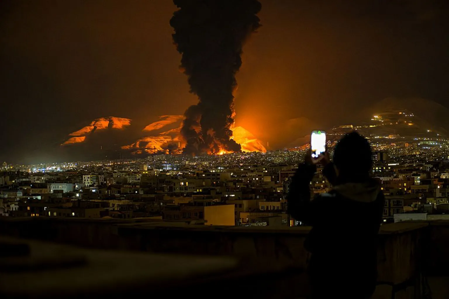  A woman uses her phone to record as smoke and flames rise at the site of airstrikes on an oil depot in Tehran on 7 March, 2026. 