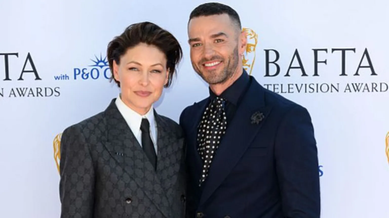 Emma and Matt Willis facing the camera, smiling. They are both wearing dark suits. Emma has cropped dark hair, and Matt has a shaved head.