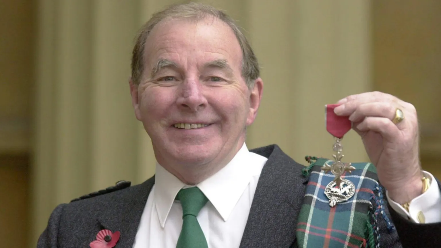  Sydney Devine smiles at the camera. He wears a kilt with a grey blazer, green tie and white shirt, with tartan wrapped around his shoulder and a red poppy pinned to his chest. He holds his MBE up for the camera. 