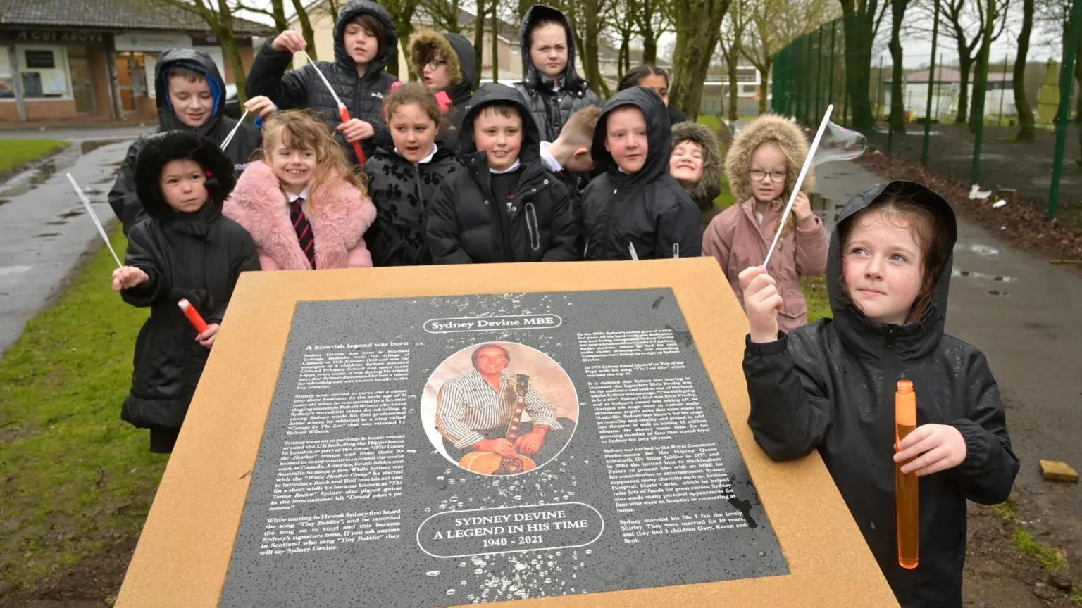 Children in waterproof clothing gather round a black memorial plaque to Sydney Devine MBE. The have tubes of soapy liquid with wands blowing bubbles in the wind