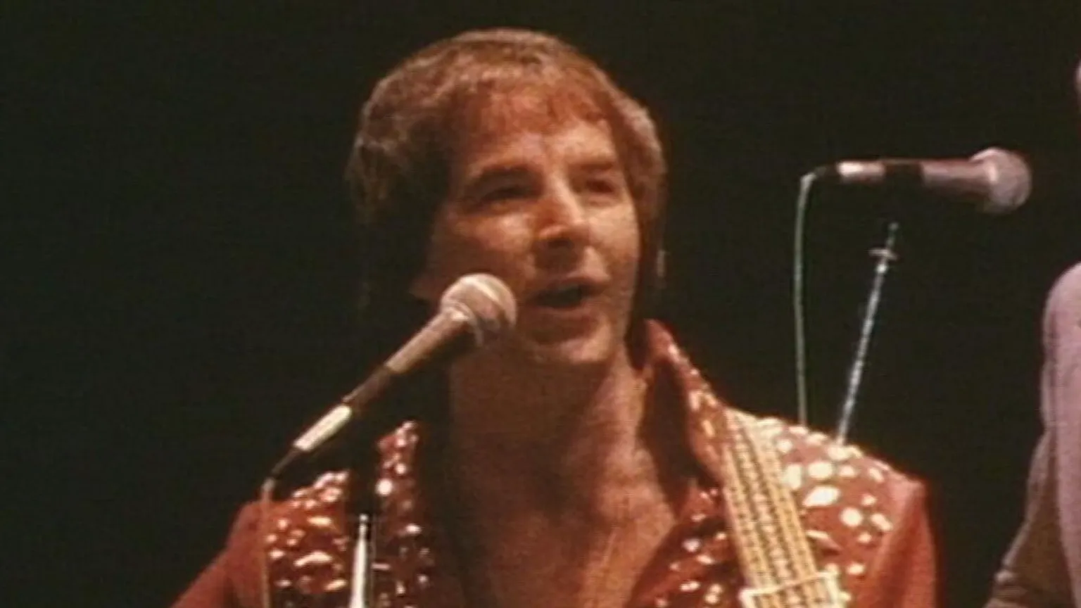 Grainy BBC archive footage of Sydney Devine who is wearing a rhinestone red body suit while strumming an acoustic guitar. A band can be seen in the shadows behind him 
