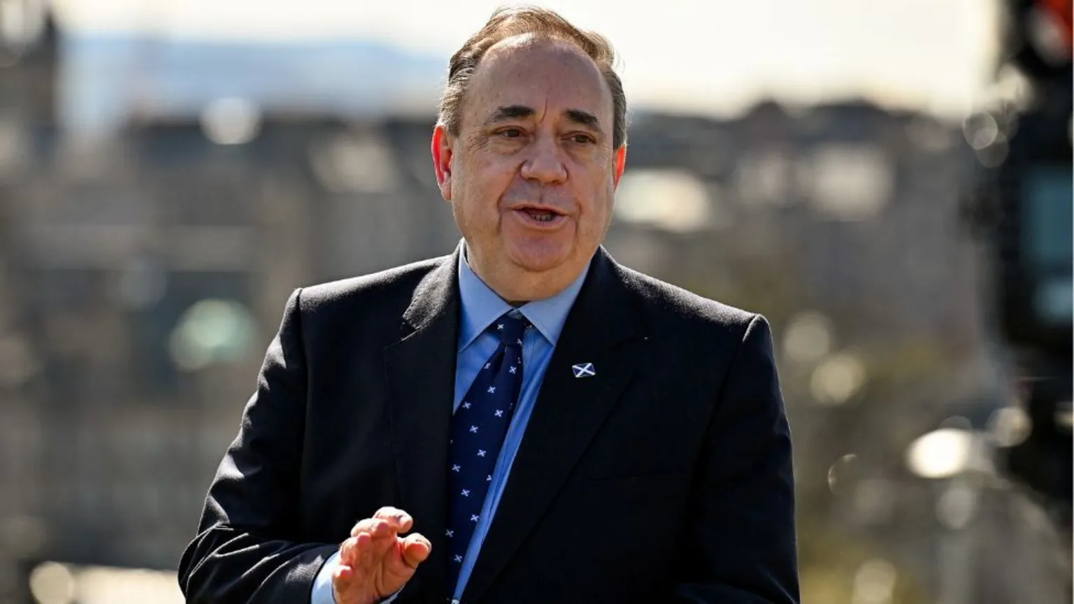 Alex Salmond, with thinning dark hair, speaks with a city backdrop behind him. He is wearing a dark suit and tie with a blue shirt.