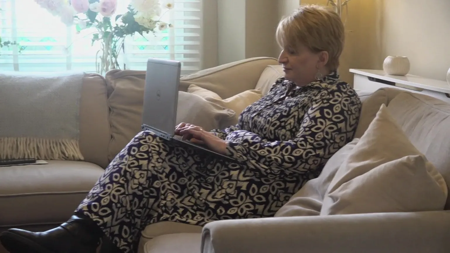 A woman with short blonde hair wearing a white and blue print maxi dress, sat on a cream corner sofa and typing on a silver laptop. In the background is a window with white blinds and a vase of flowers.