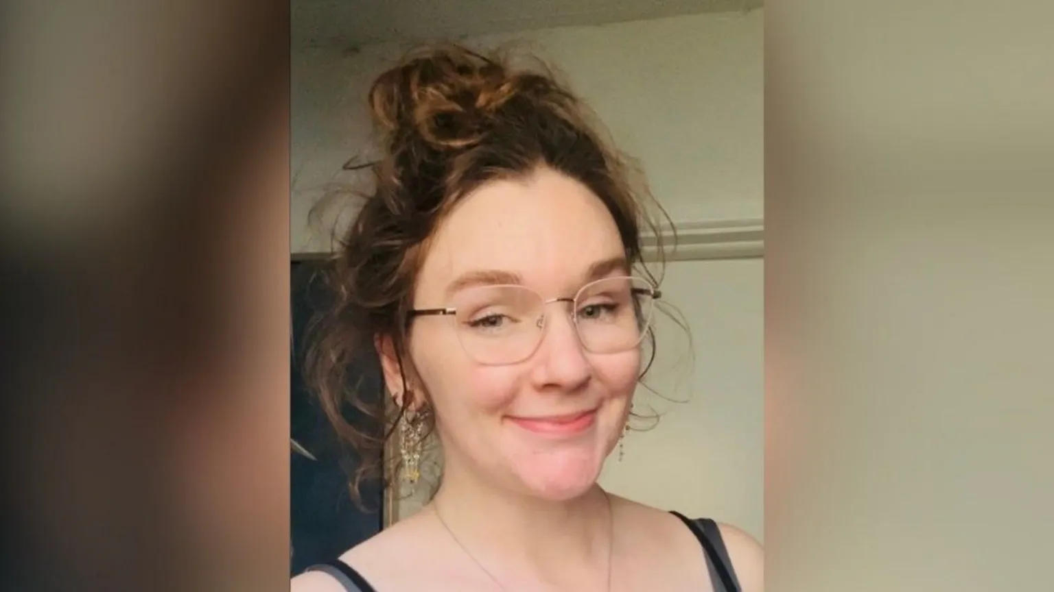 Nadia McVeigh A woman with curly brown and gold hair piled on top of her head. She has gold rimmed glasses, dangly earrings and wears a black vest top.