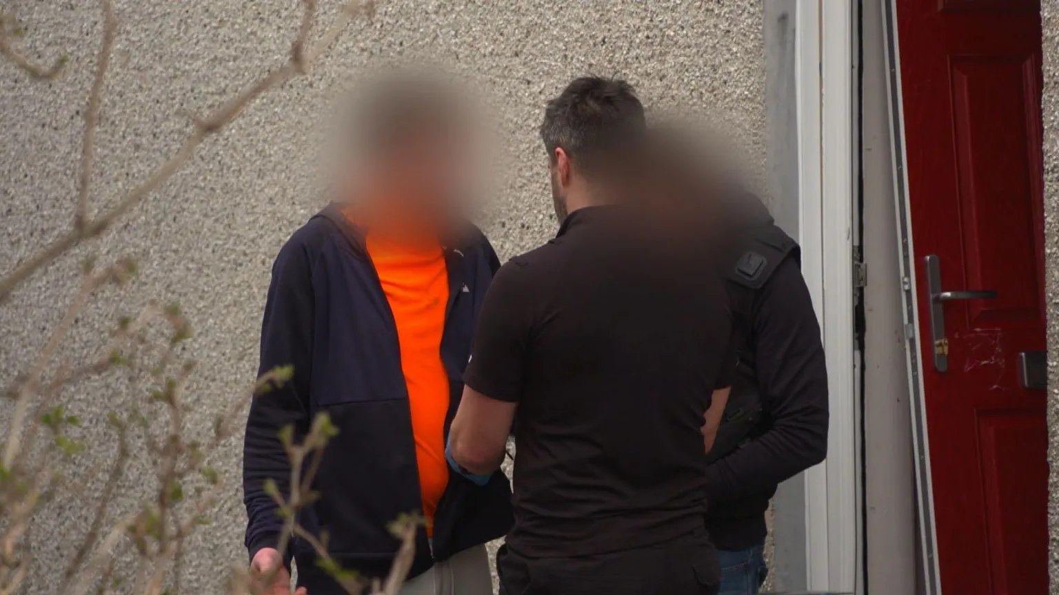 A man in a dark blue hoodie and orange t-shirt - his face blurred - is facing two police officers - both of whom are plainclothes and turned away from the camera. To the right, a red door lies off its hinges in a pebble dashed wall.
