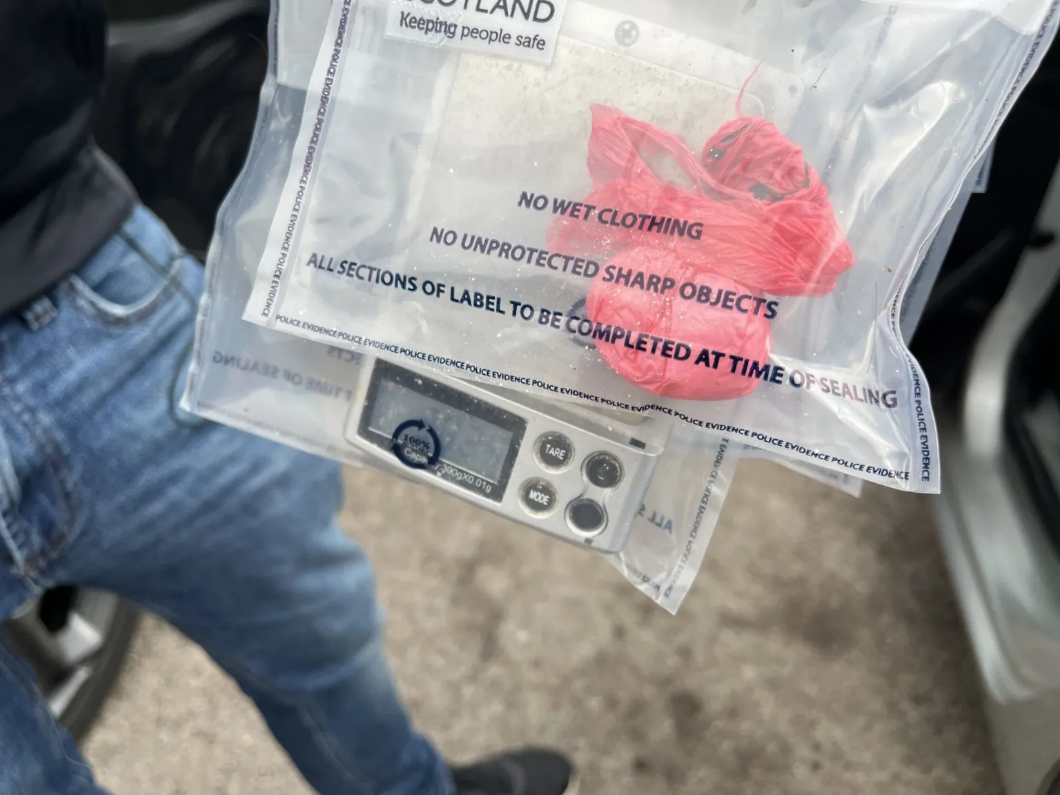Photo of two police evidence bags being held by someone in blue jeans. One bag contains a red wrapper with white powder; the other has a set of digital scales.