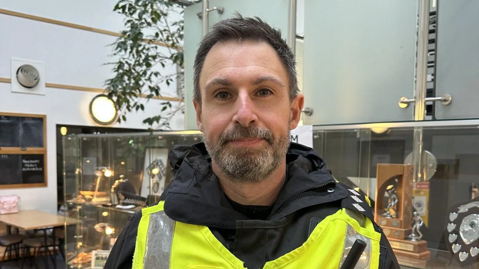 A man in his mid 40s wearing a hi-vis police vest and dark uniform looks at the camera. He has short dark hair and a short greying beard.
