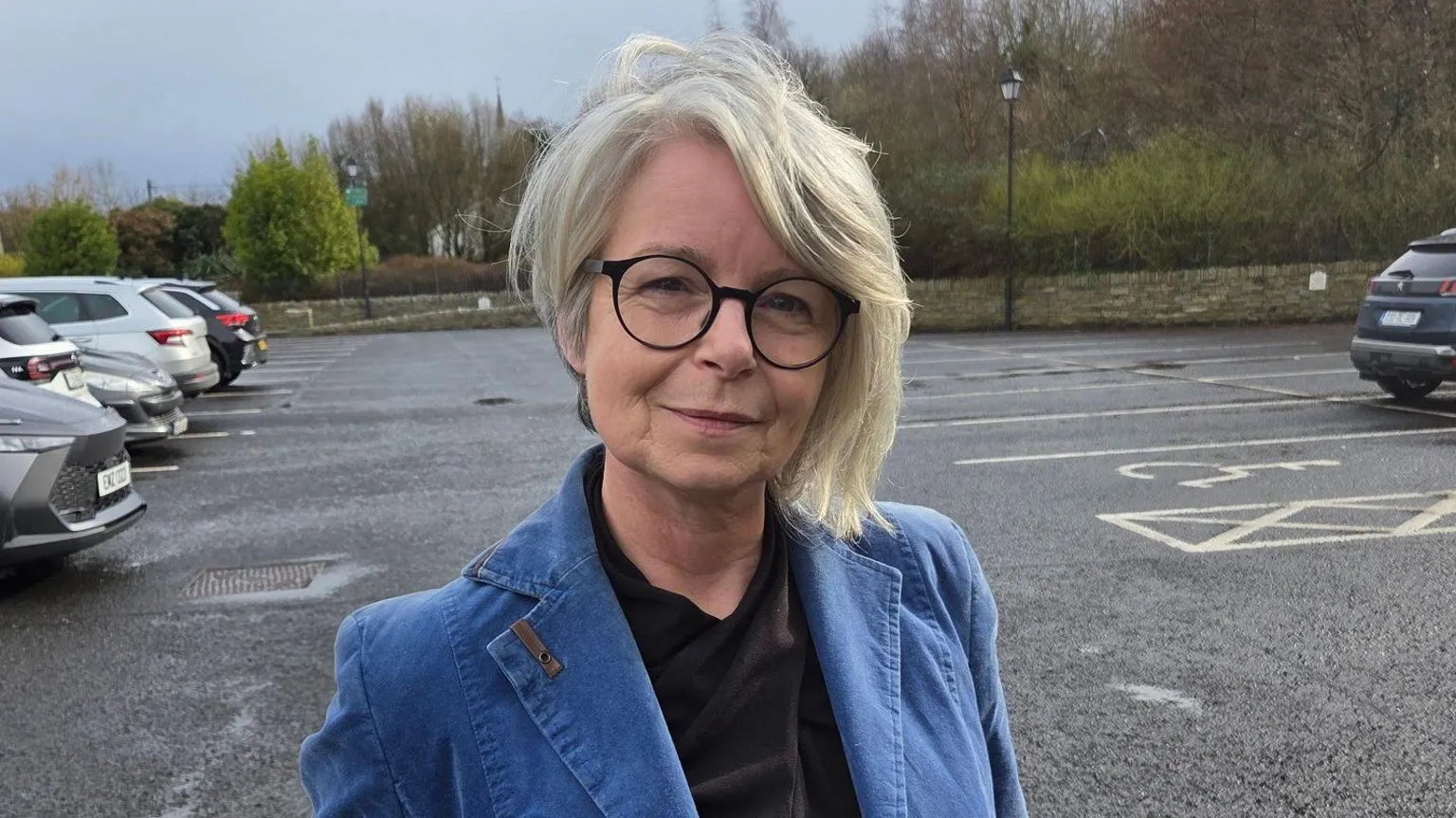 A lady with shoulder lenght fair hair stands in a car park. she is wearing a blue jacket and black blouse. She is wearing dark rimmed glasses.