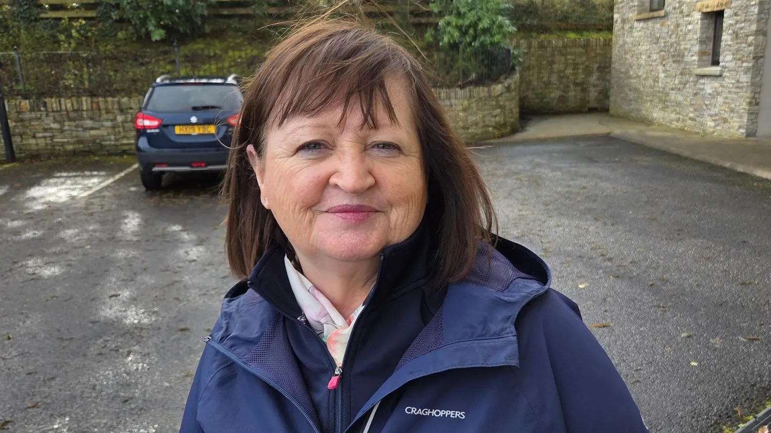 A lady in a navy blue rain coat stands in front of a parked car. The walls of a house are seen in the background. She has shoulder length brown hair.