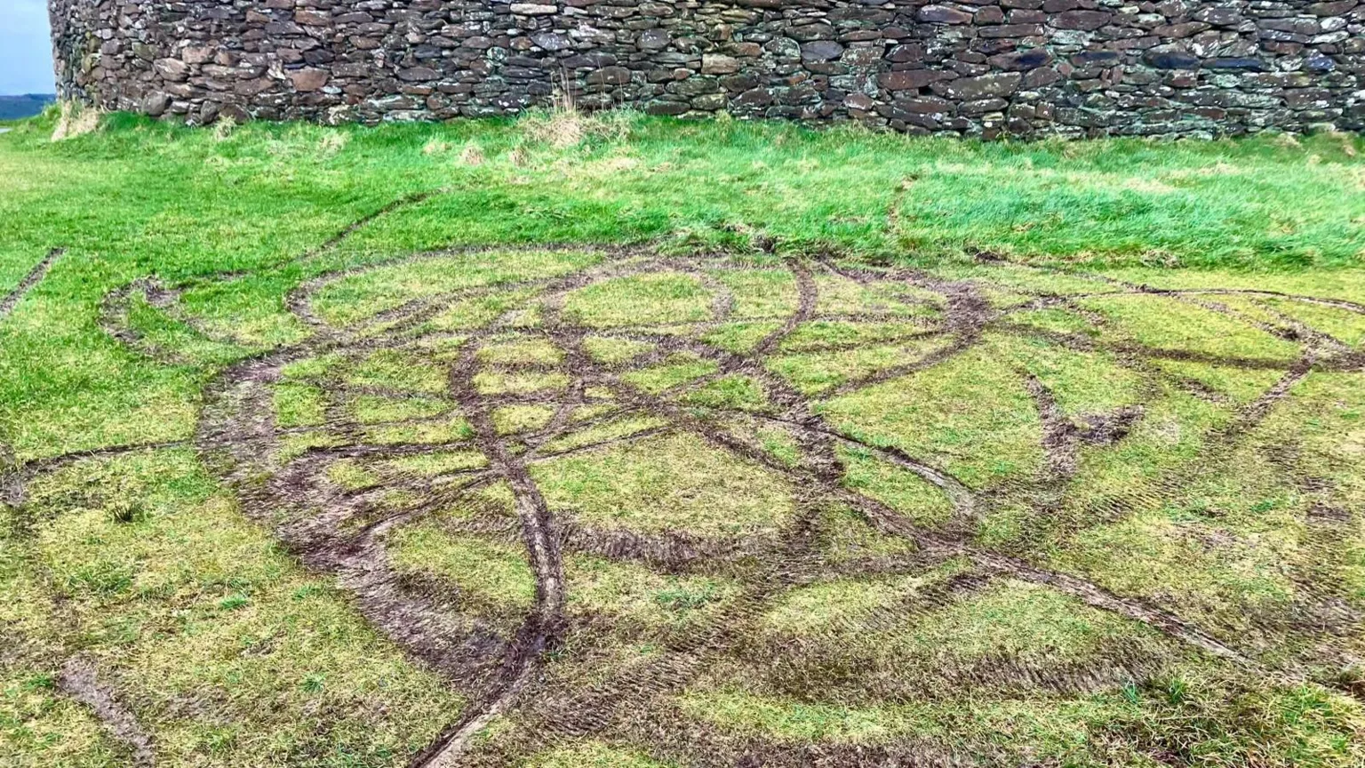 grass ripped up by tyre tracks is in the foreground while the stone walls of a fort are in the background