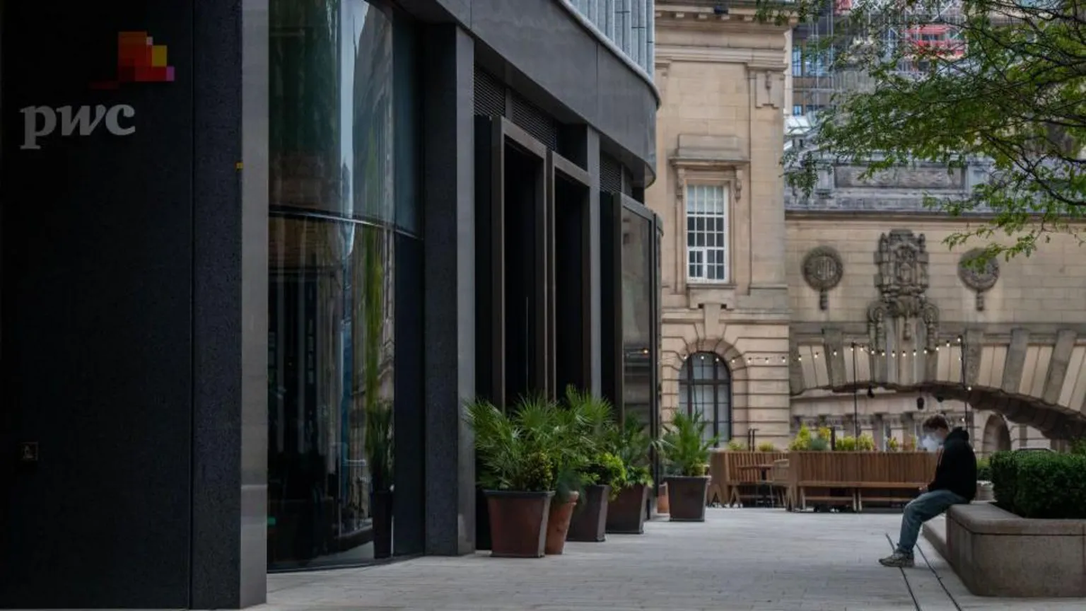 Bloomberg via Outside of a shiny dark building seen from an angle carring the PwC logo. In the background is an older stone building and a young man in the middle distance sitting on a wall vaping.