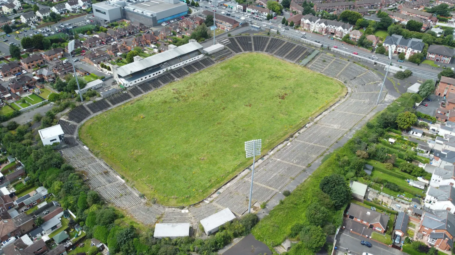 An aerial view of a grass pitch with concrete terrace-style stands. It sits in a residential area with a number of red brick houses surrounding the site.