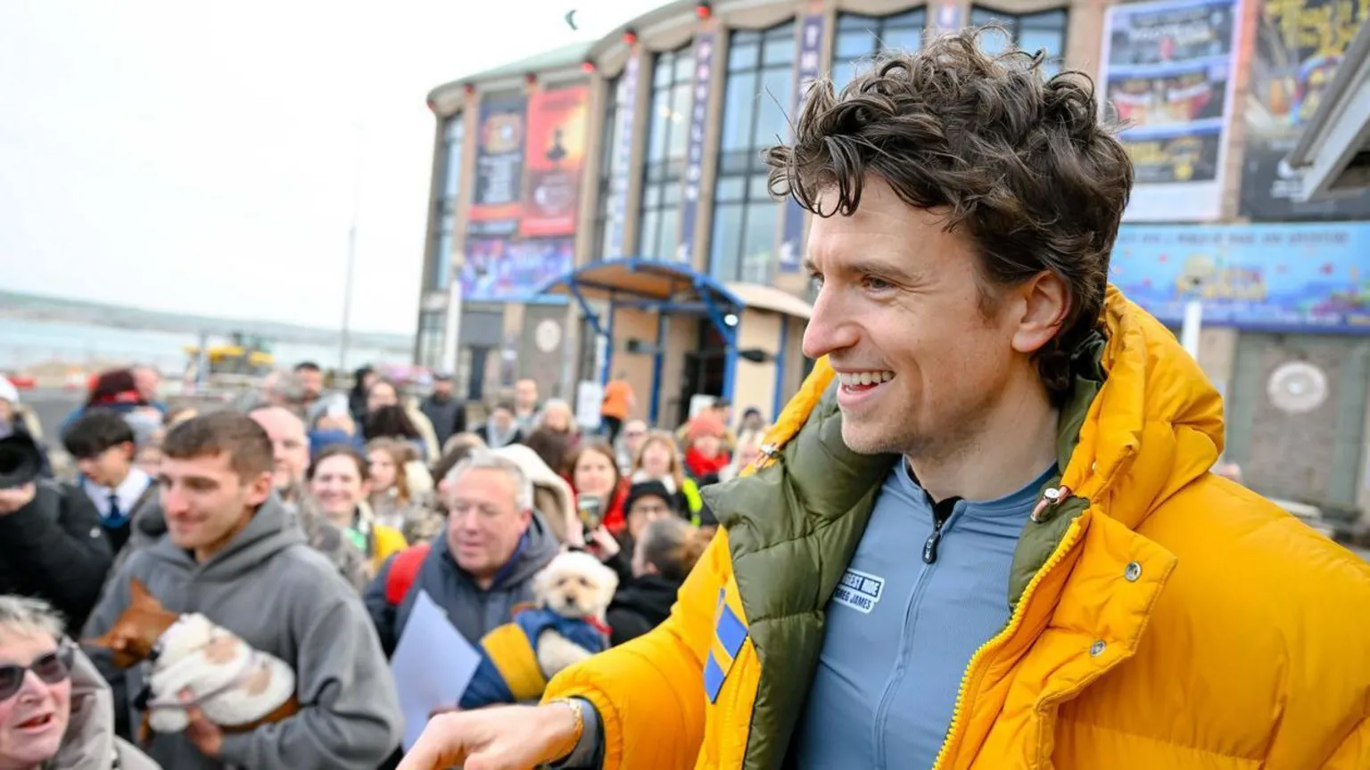 Greg James smiling at the camera in a close-up photo . He's a white man with curly brunette hair. He's wearing a bright yellow puffer coat open with a light grey blue cycling top underneath. There are crowds of people behind him, he's smiling at them.