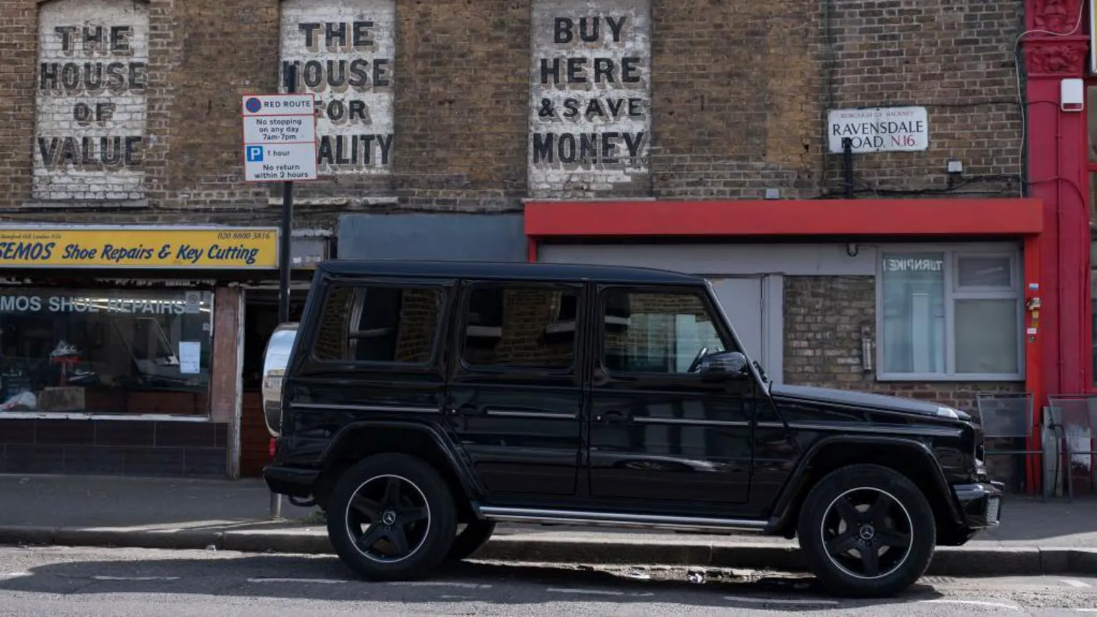  A large black SUV is parked outside shops on Ravensdale Road in N36, including a shoe repair and key cutting business. There are Victorian style markings on the brickwork above the shops, advertising businesses that used to be there