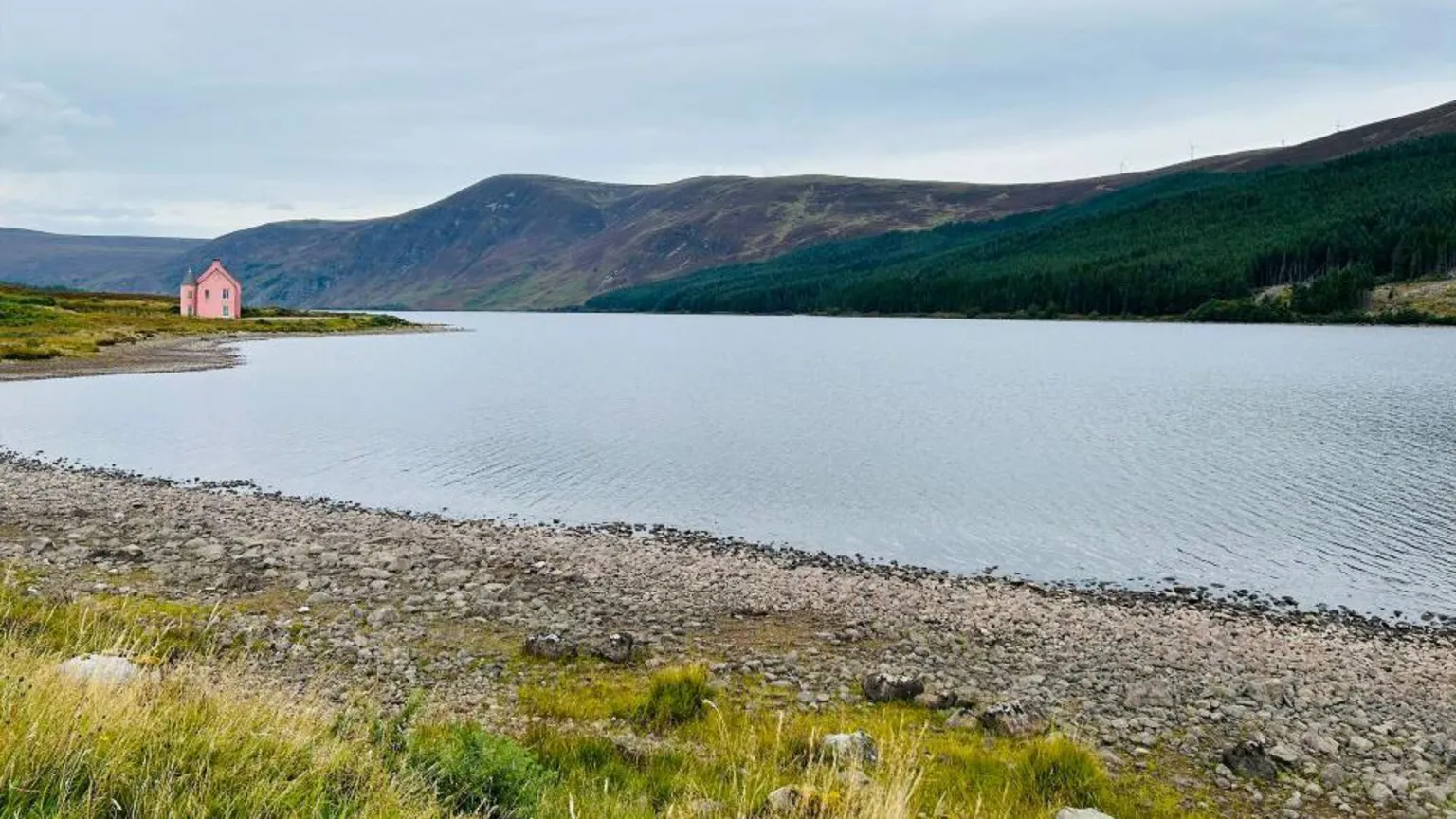 Roseanna Mitchell A small pink house sits beside a wide lake surrounded by rolling hills and dark pine forests under an overcast sky.