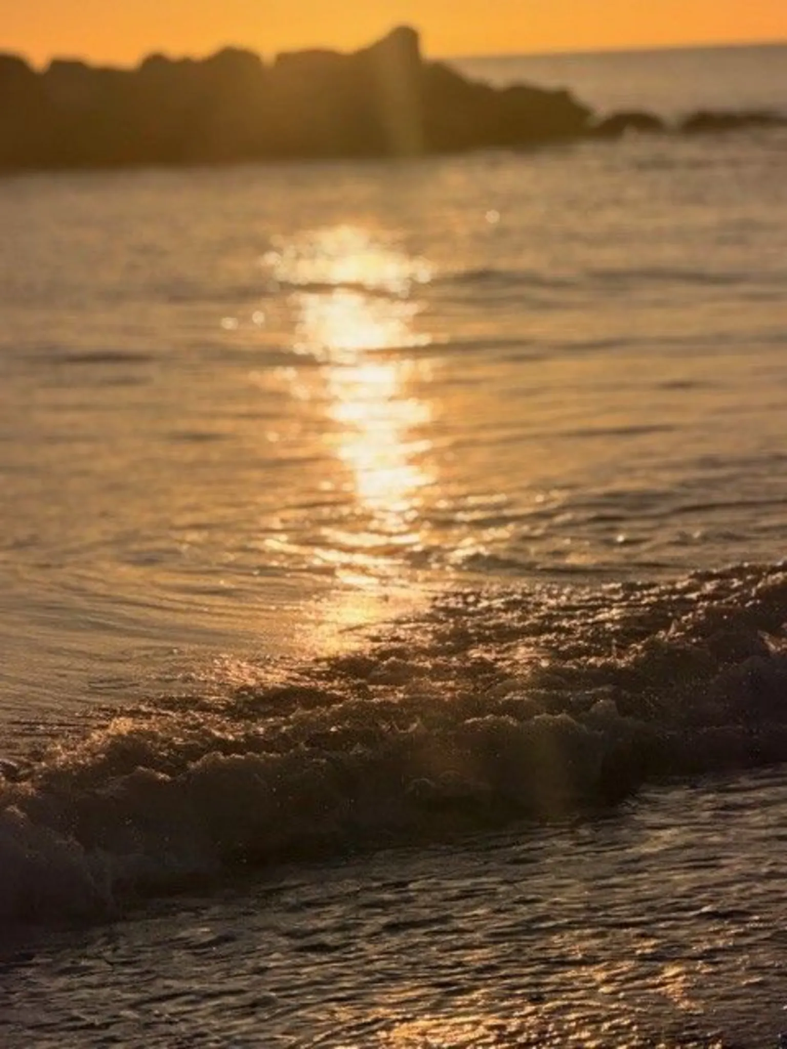 Janice Mitchell Golden sunlight reflecting on gentle ocean waves near the shore, with dark rocks silhouetted in the background at sunset.