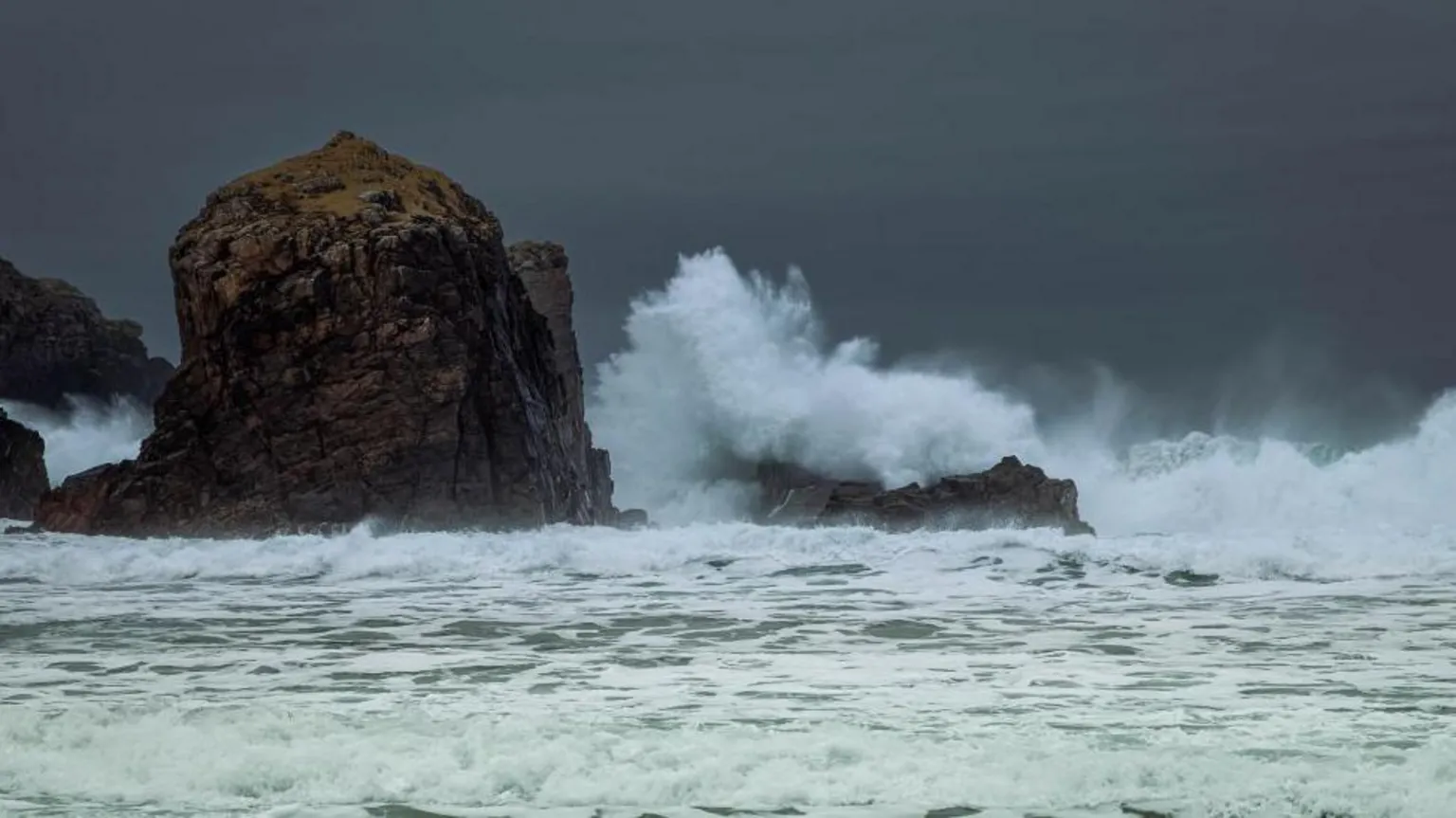 Andrew Briggs Large waves crash dramatically against dark, jagged coastal rocks under a stormy, grey sky.