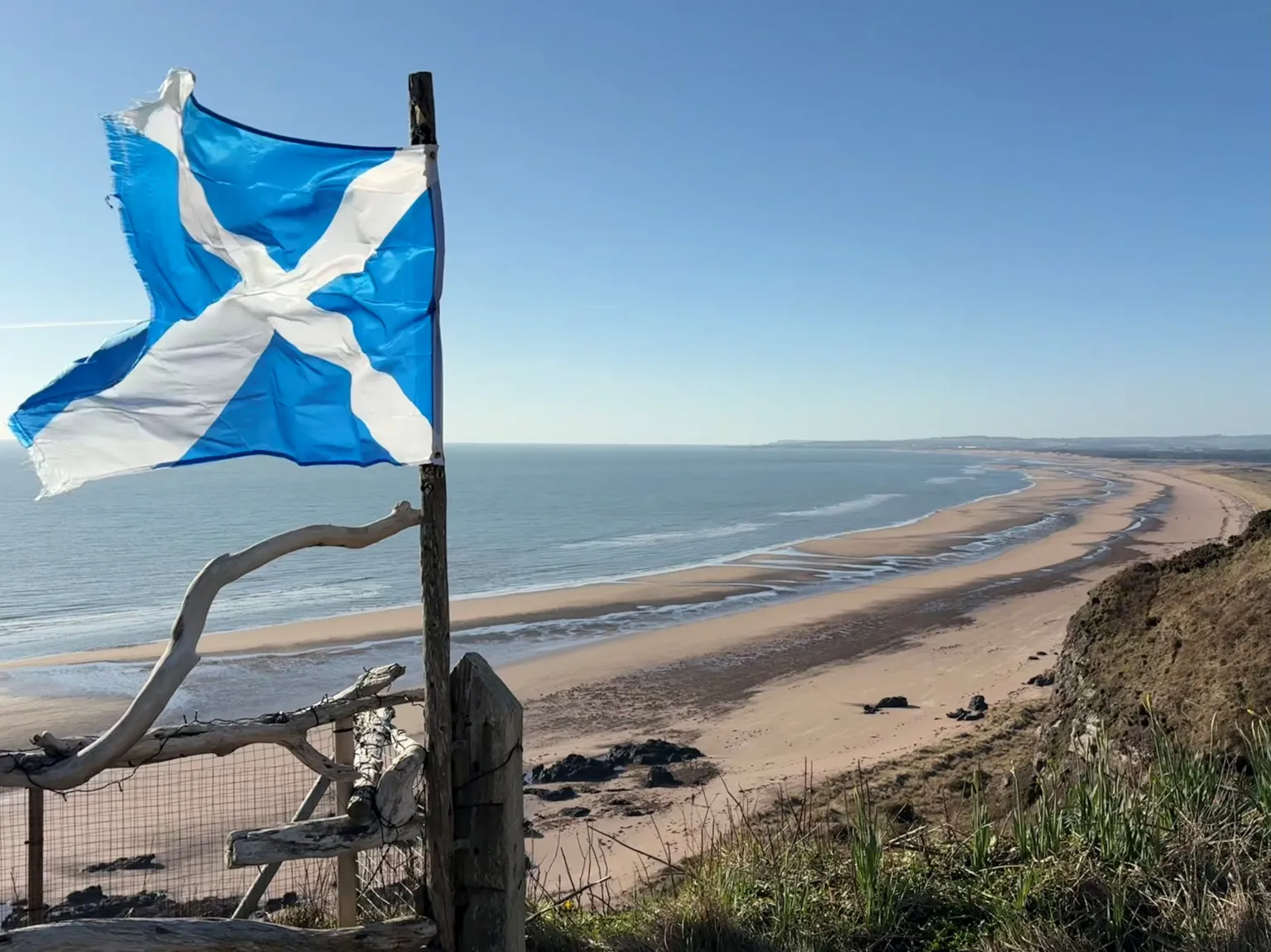 Alison Cameron A tattered blue-and-white Saltire flag flutters on a wooden post overlooking a long, sandy beach and calm sea under a clear sky.