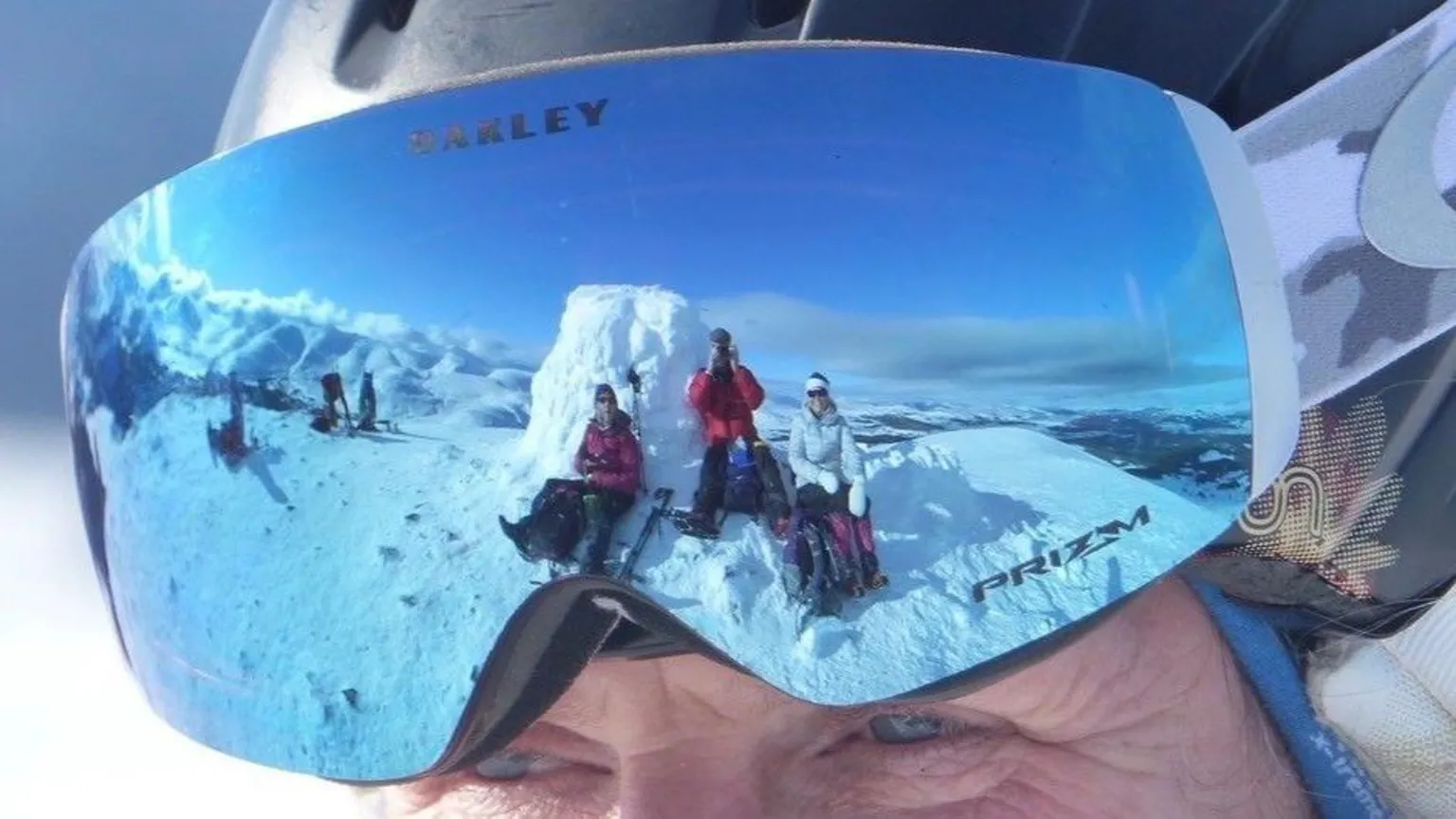 Richard Castro A snowy mountain landscape reflected in the mirrored ski goggles of a person wearing a helmet, showing several people sitting on the snow with wide views of peaks and sky.