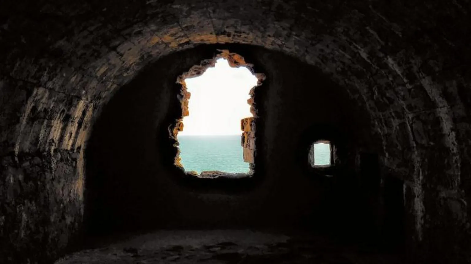 Aaren Dannoch A dark stone tunnel with a rough arched ceiling opens onto a bright view of the sea through a jagged, irregular window in the far wall.