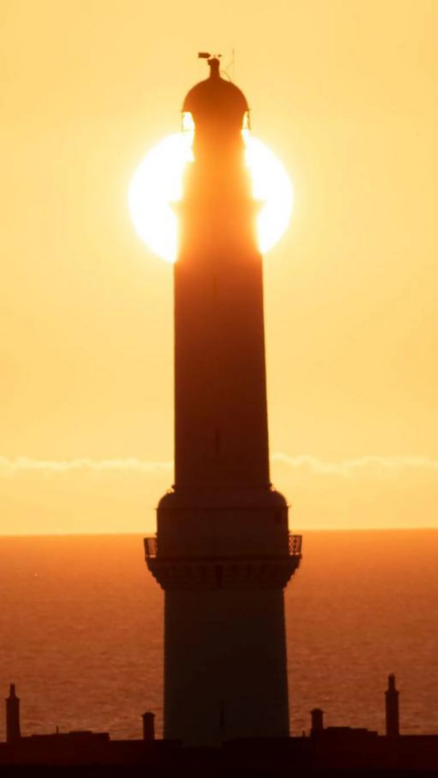 Lee Shirreffs A lighthouse in silhouette stands directly in front of the rising or setting sun, creating a glowing halo around its tower above the sea.