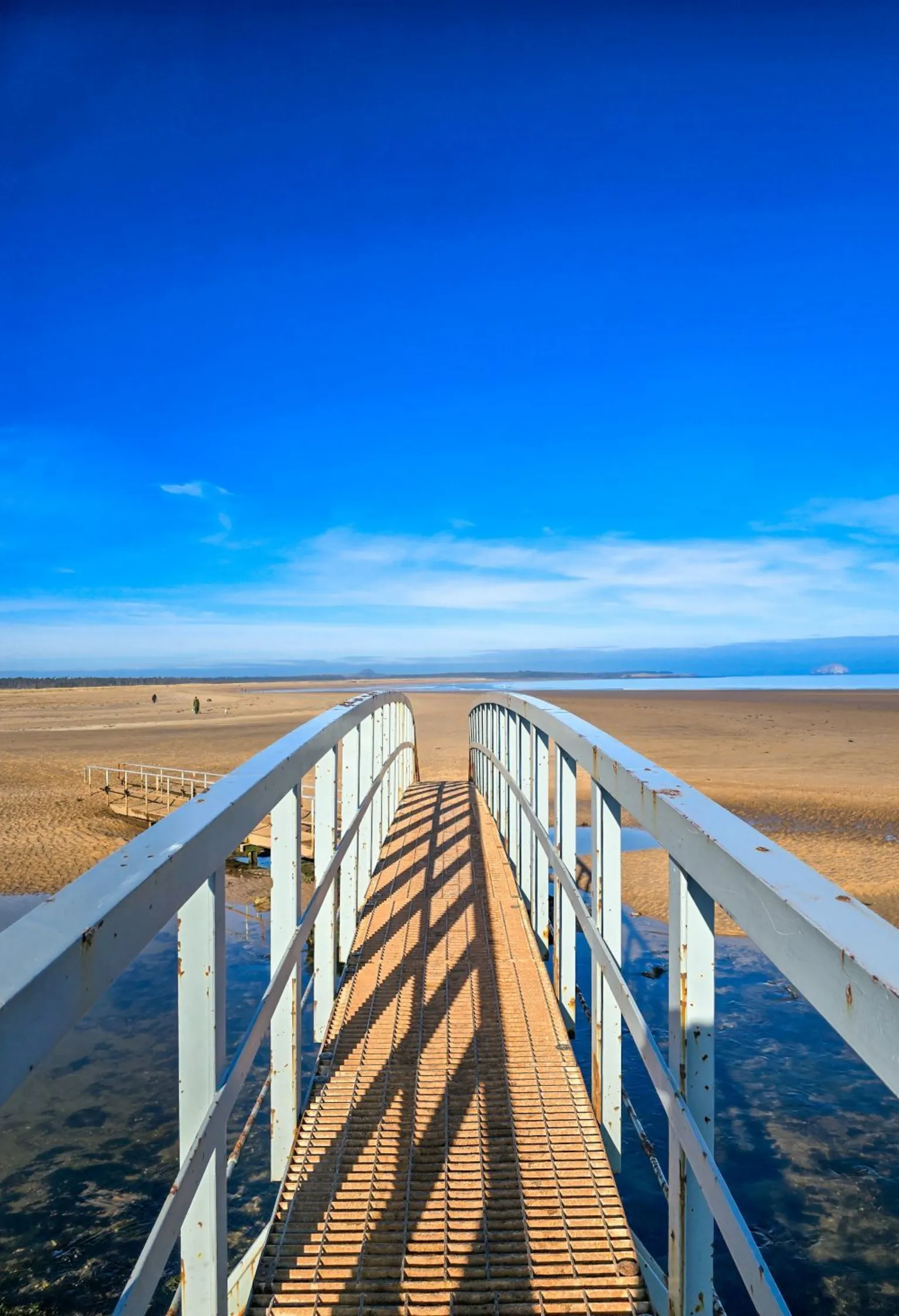 Nicola Orr A white metal footbridge extends over shallow water toward a wide, sandy beach under a bright blue sky.
