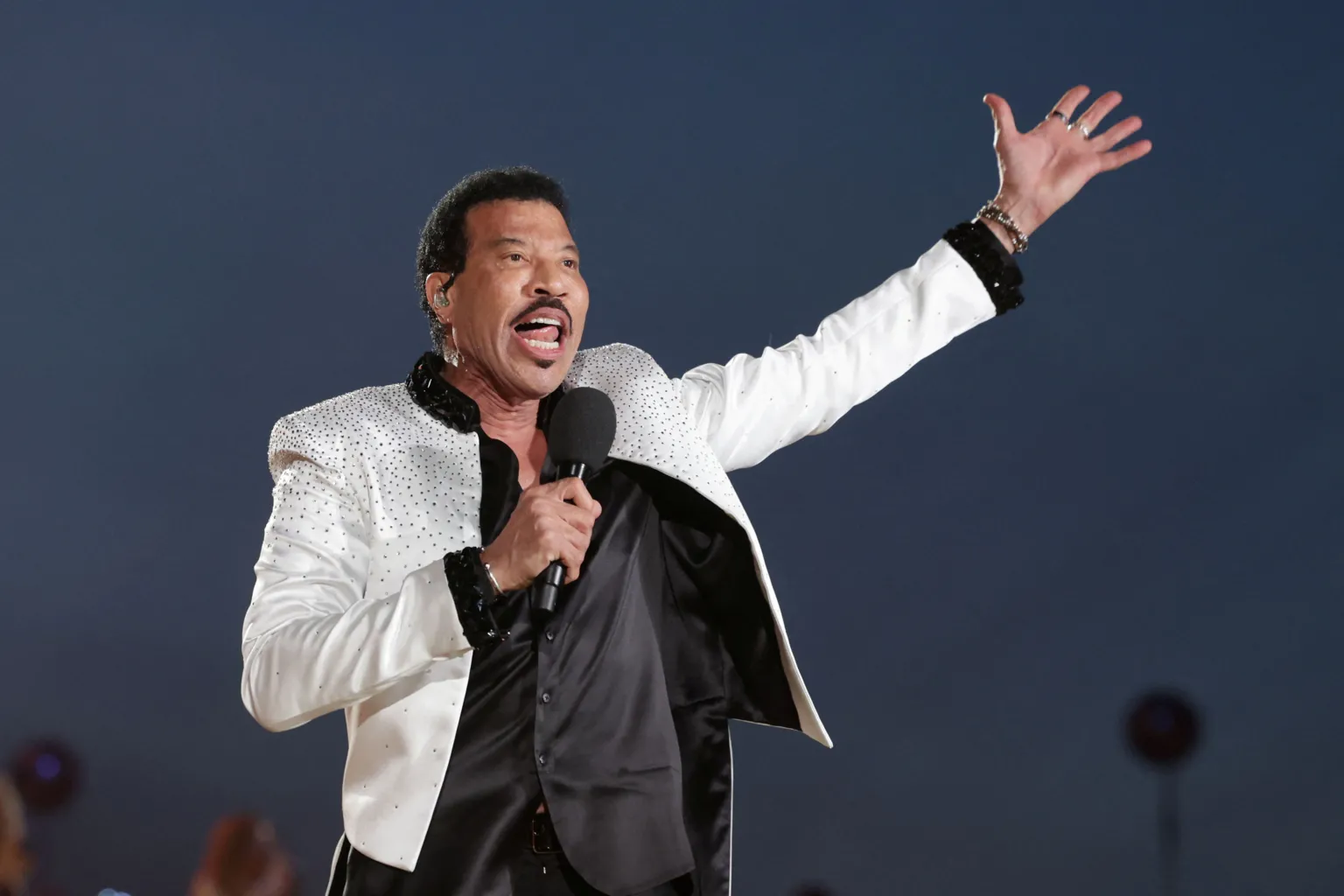 Lionel Richie in a white, jewel-encrusted jacket, raises a hand to the air as he performs in concert