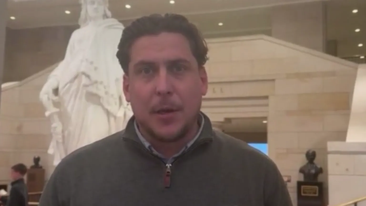 Joe Bennett A man with brown hair stands inside the McCain Institute in Washington. He is wearing a great 1/4 zip jumper and a name badge that reads 'Joe Bennett'. There is a grand marble staircase in the background and a statue. 