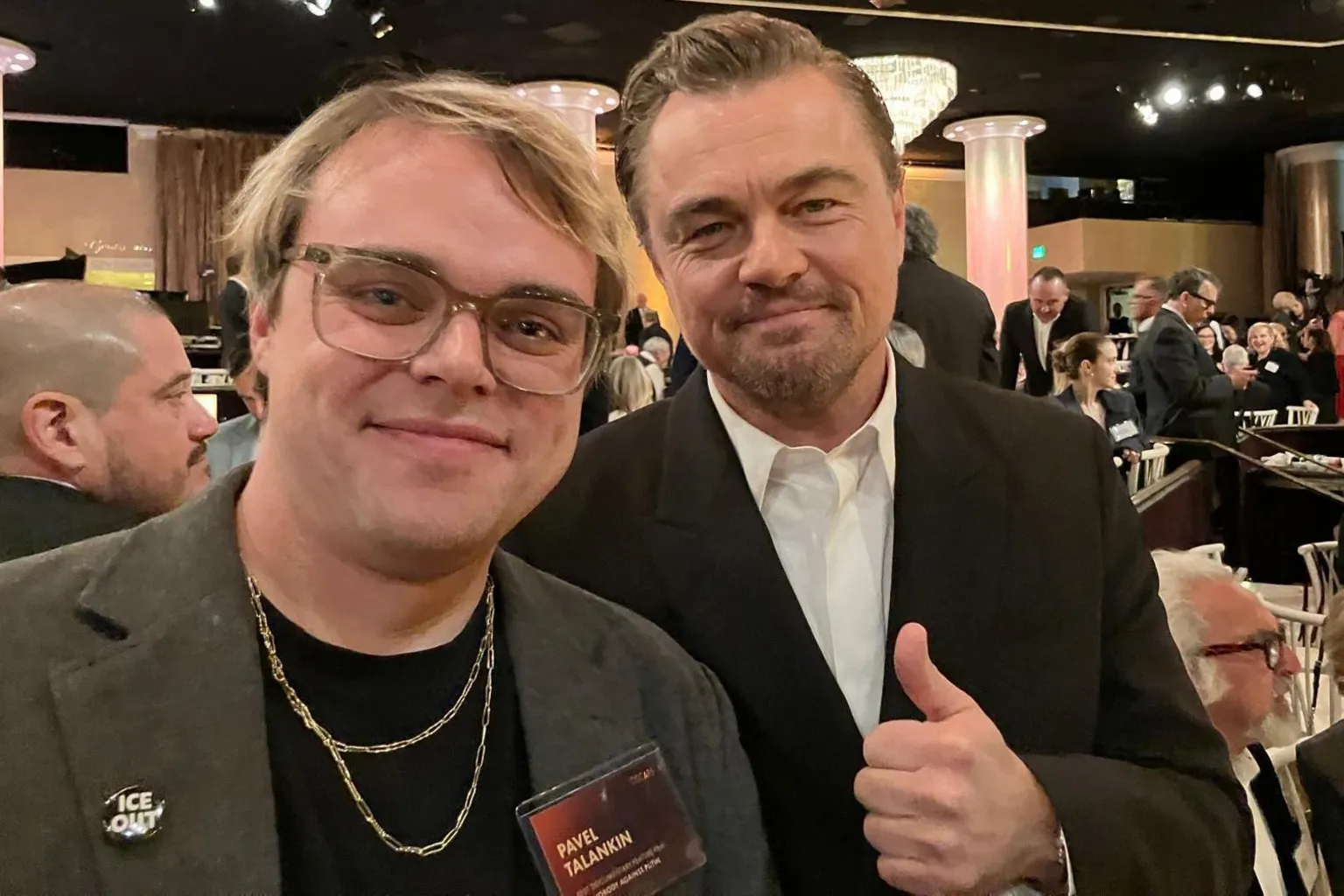 Pavel Talankin On the left - Pavel Talankin smiling, with glasses, khaki jacket & black top & gold chain On the right -- Leonardo DiCaprio, smilnig, dark jacket, white shirt, giving a thumbs up