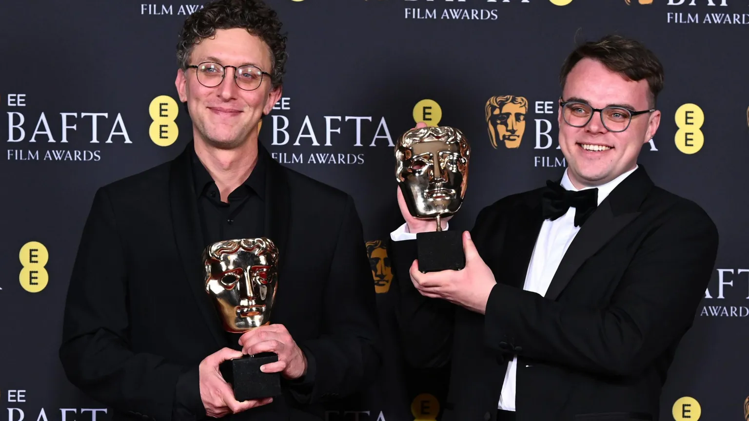 David Borenstein on the left. Dark curly hair, glasses, smiling. Black suit. and Pavel Talankin on the right. brown hair, glasses, wide smile. Black dinner jacket and bow tie. Both holding the Documentary Award for 'Mr Nobody Against Putin' during the BAFTA Film Awards in February. Background is black with BAFTA logo and words EE BAFTA Film Awards