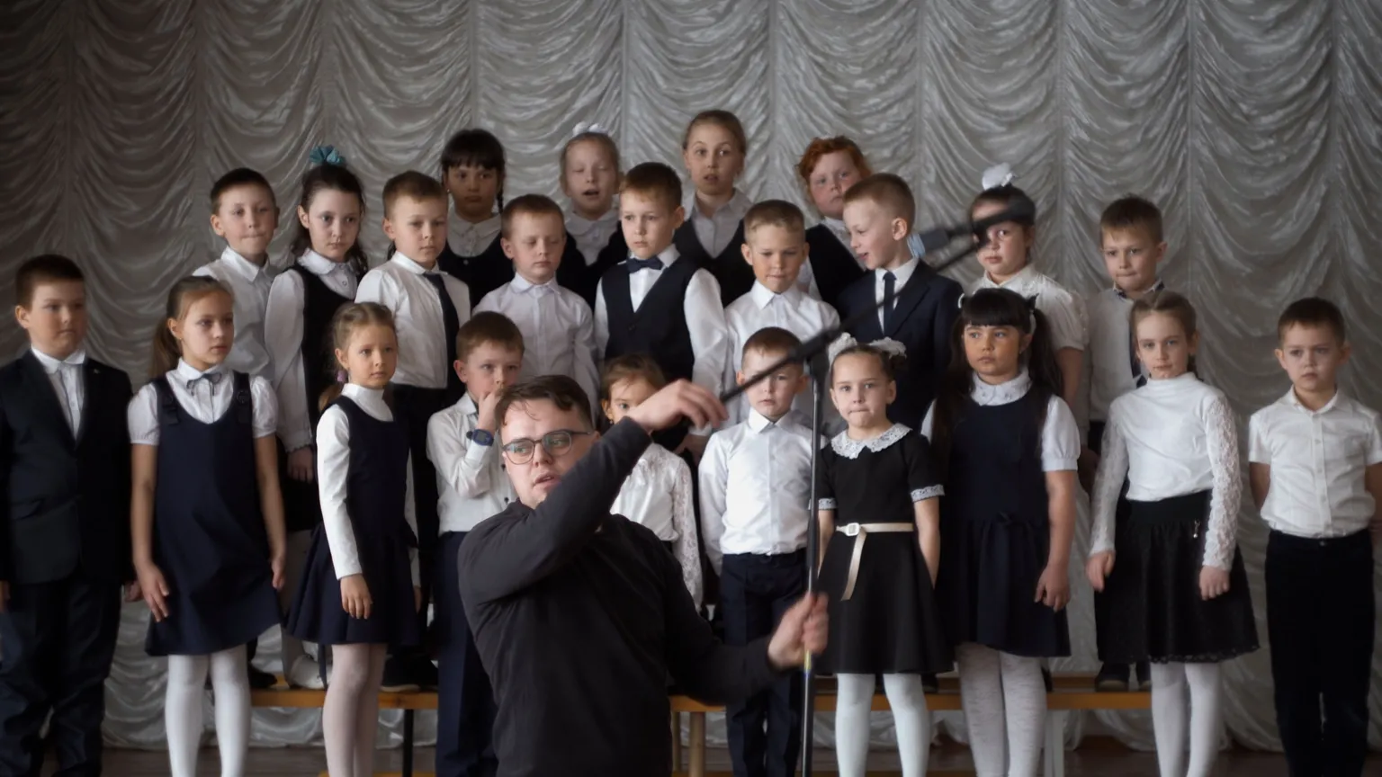 Pavel Talankin School choir -- girls in white tops and black dress or skirt, boys in white shirt and trousers, one boy with black jacket, one boy in waistcoat and bow tie Pavel Talankin in the bottom centre in black setting up microphone
